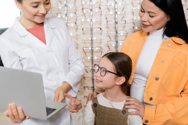 Smiling girl in eyeglasses pointing at laptop near oculist and asian mother in optics salon — Stock Photo