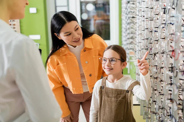 Positive kid choosing eyeglasses near asian mom and blurred ophthalmologist in optics shop - foto de stock