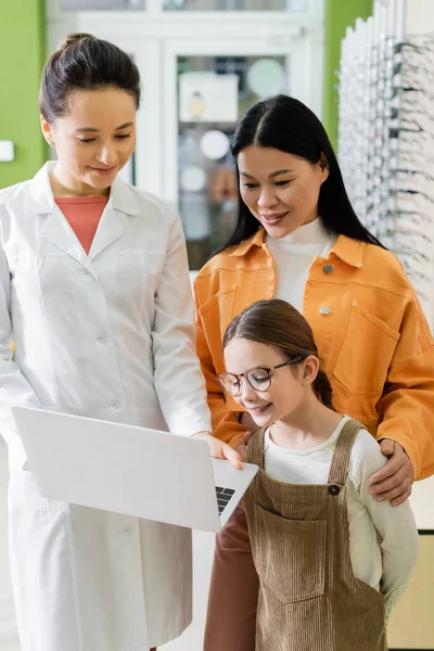 Chica en gafas mirando portátil cerca de asiático mamá y sonriente oculista en óptica tienda - foto de stock