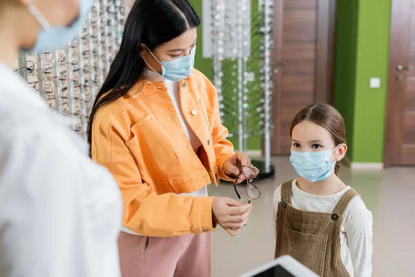 Asian woman in medical mask holding eyeglasses near daughter and blurred oculist in optics store - foto de stock