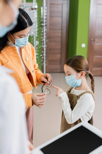 Дівчина в медичній масці, вказуючи на окуляри в руках азіатської матері і розмитого окуліста в салоні оптики — стокове фото