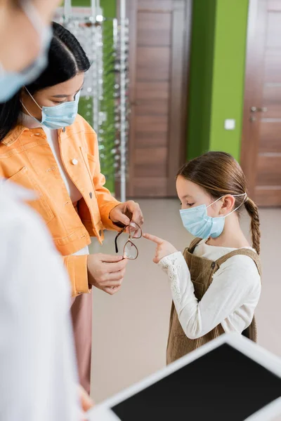 Girl in medical mask pointing at eyeglasses in hands of asian mother and blurred oculist in optics salon — Stock Photo