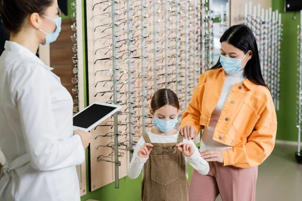 Girl in medical mask choosing eyeglasses near asian mother and blurred oculist with digital tablet — Photo de stock