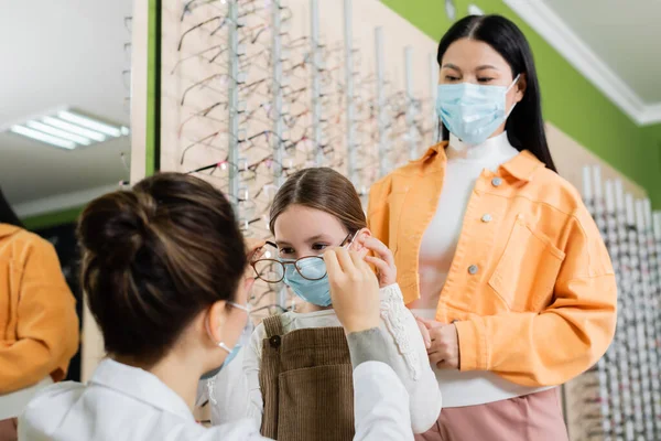 Blurred ophthalmologist trying eyeglasses on girl in medical mask near asian woman in optics store — стоковое фото