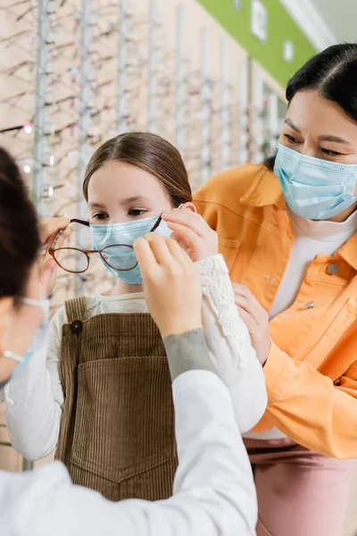 Blurred oculist trying eyeglasses on girl in medical mask near asian mother in optics salon — Stock Photo