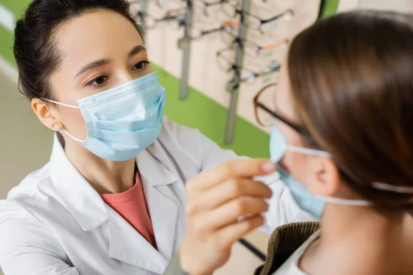 Oculist in medical mask trying eyeglasses on blurred girl in optics store — стоковое фото