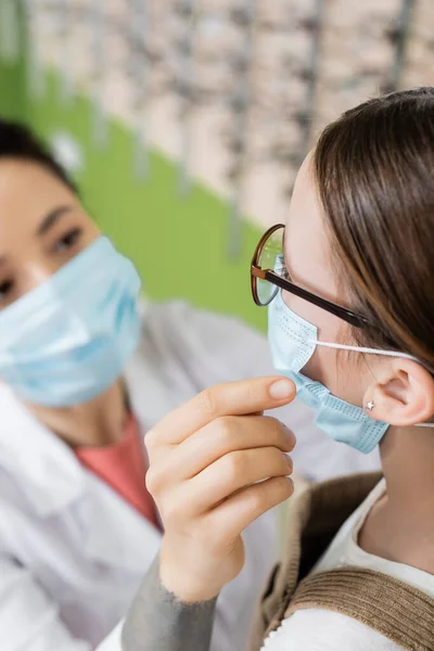 Розмитий окуліст в медичній масці біля дівчини в окулярах в салоні оптики — стокове фото