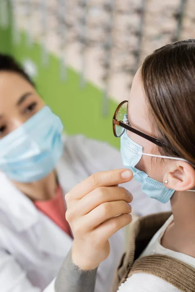 Blurred oculist in medical mask near girl in eyeglasses in optics salon — Stock Photo