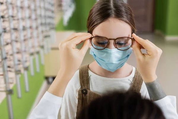 Blurred ophthalmologist trying eyeglasses on girl in medical mask in optics salon — Stock Photo