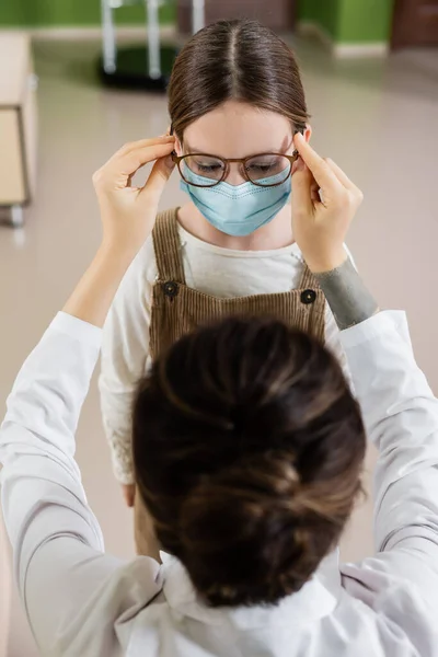 Blurred ophthalmologist trying eyeglasses on child in medical mask in optics store - foto de stock