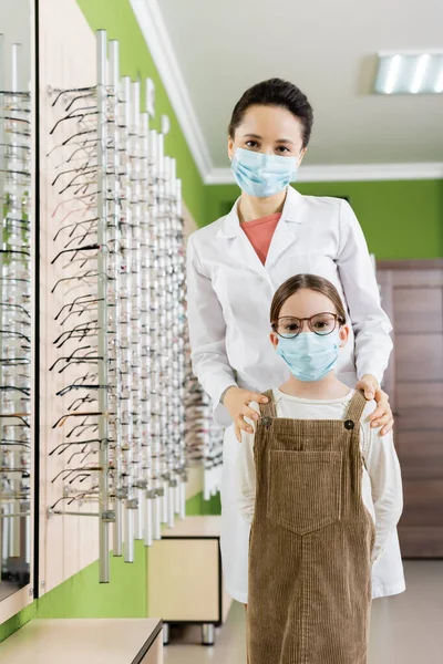 Oculist and girl in medical masks looking at camera near assortment of eyeglasses in optics store - foto de stock