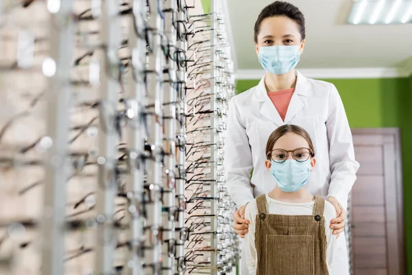 Ophthalmologist and girl in medical masks looking at camera near assortment of eyeglasses in optics store on blurred foreground — Stock Photo