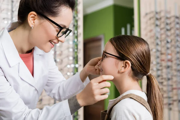 Side view of smiling ophthalmologist trying eyeglasses on child in optics salon — Stock Photo