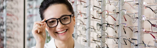 Smiling oculist adjusting eyeglasses while looking at camera in optics shop, banner — Stock Photo