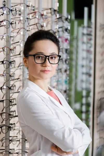 Oculist in white coat standing with crossed arms near assortment of eyeglasses in optics shop — Stockfoto