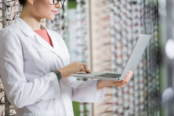 Cropped view of oculist in white coat using laptop in optics shop on blurred background — Stock Photo