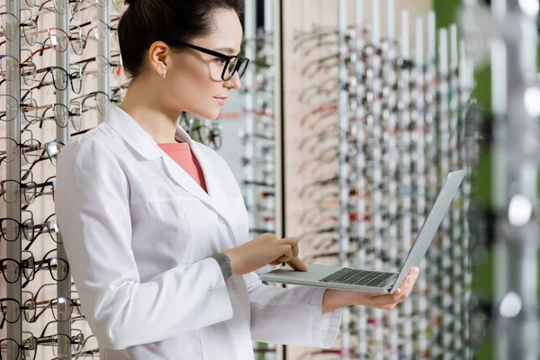 Young oculist using laptop near assortment of eyeglasses in optics store — Fotografia de Stock