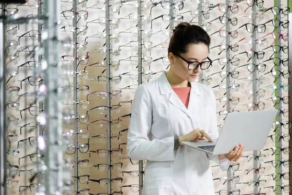 Oculist in eyeglasses and white coat using laptop while working in optics store - foto de stock