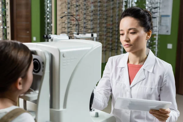 Ophthalmologist with digital tablet examining vision of kid on autorefractor in optics salon — Fotografia de Stock