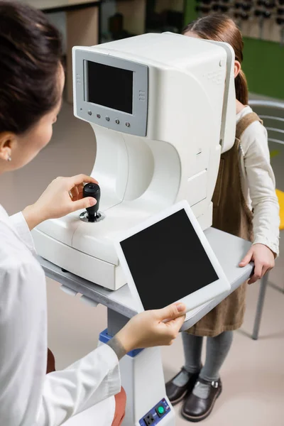 Blurred ophthalmologist holding digital tablet with blank screen while measuring eyesight of girl on autorefractor in optics store — стоковое фото