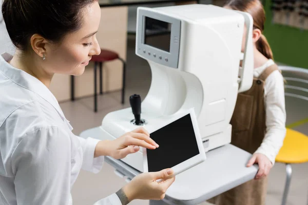 Oculista sonriente usando tableta digital con pantalla en blanco mientras mide la visión de la chica borrosa en el oftalmoscopio - foto de stock