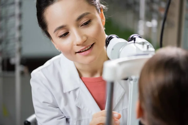 Positive optometrist checking eyesight of girl on autorefractor in optics store - foto de stock