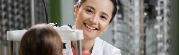 Happy oculist looking at camera while measuring vision of girl on ophthalmoscope in optics store, banner — Stockfoto