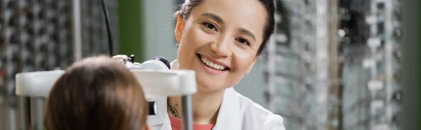 Happy oculist looking at camera while measuring vision of girl on ophthalmoscope in optics store, banner — Fotografia de Stock