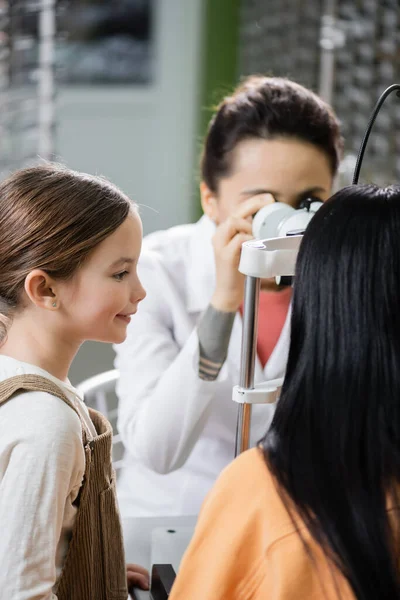 Blurred optometrist checking eyesight of woman on vision screener near smiling girl in optics store — Photo de stock