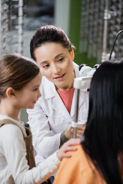 Positive optometrist measuring eyesight of woman on autorefractor near girl in optics store — Fotografia de Stock