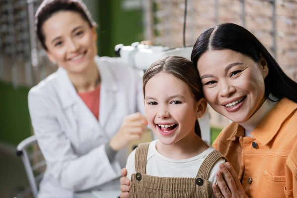 Excited interracial mother and daughter looking at camera near blurred oculist in optics store — Stockfoto