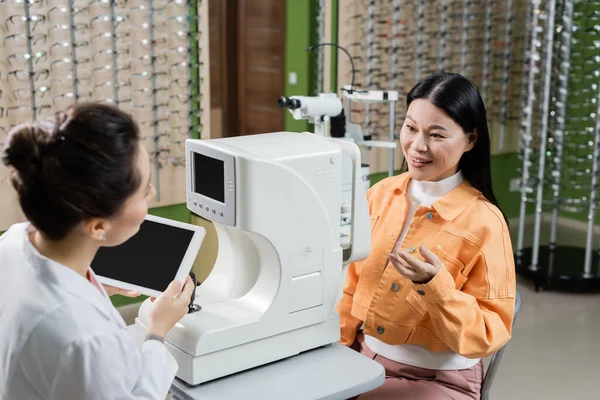 Souriant asiatique femme pointant avec main près flou oculiste avec tablette numérique et visionneuse dans l'optique boutique — Photo de stock