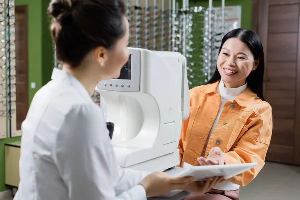 Smiling asian woman pointing at digital tablet near blurred oculist and autorefractor in optics salon - foto de stock