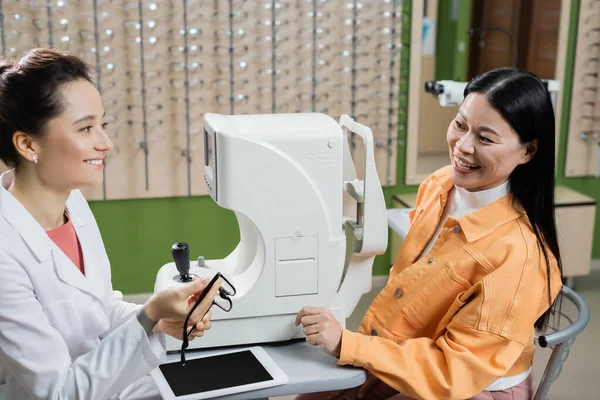 Sonriente optometrista celebración gafas cerca feliz asiático mujer y oftalmoscopio en óptica tienda - foto de stock