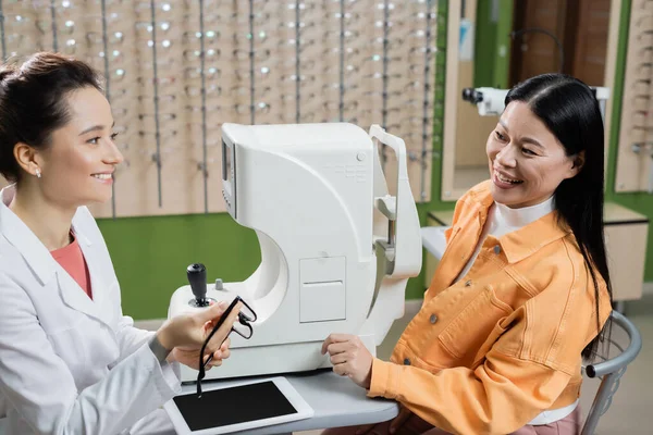 Smiling optometrist holding eyeglasses near happy asian woman and ophthalmoscope in optics shop — стоковое фото