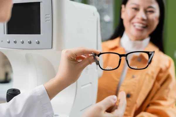 Selective focus of eyeglasses in hand on oculist near vision screener and smiling asian woman — Foto stock