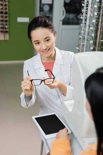 Oftalmólogo sonriente sosteniendo anteojos cerca de una mujer borrosa, tableta digital y proyector de visión en la tienda de óptica - foto de stock