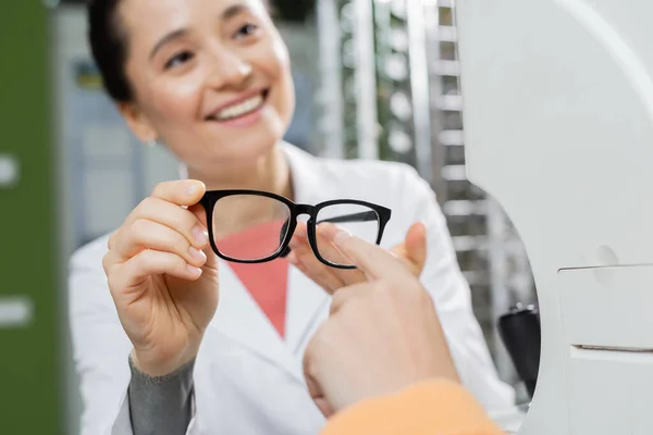 Woman pointing with finger at eyeglasses in hand of happy oculist in optics shop — Foto stock