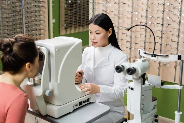 Asian optometrist checking vision of blurred woman on ophthalmoscope in optics store — Fotografia de Stock