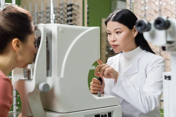 Asian oculist pointing at vision screener while measuring eyesight of blurred woman in optics store — Fotografia de Stock