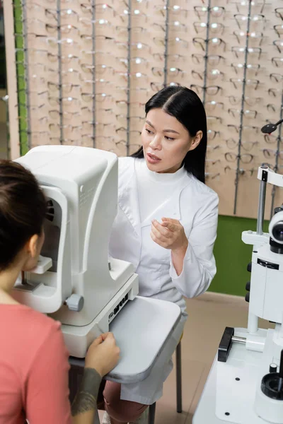 Ophtalmologiste asiatique pointant avec le doigt tout en mesurant la vision de la femme floue sur autorefractor — Photo de stock
