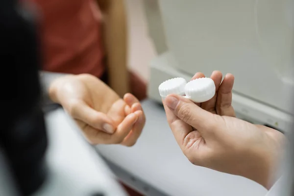 Partial view of ophthalmologist giving lens case to woman on blurred foreground - foto de stock