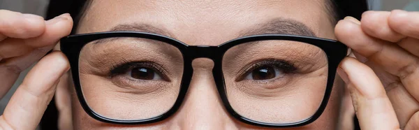 Partial view of asian woman adjusting eyeglasses while looking at camera, eye care concept, banner — Stock Photo