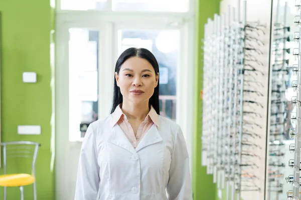 Asian oculist in white coat looking at camera near blurred assortment of eyeglasses in optics salon — Stock Photo