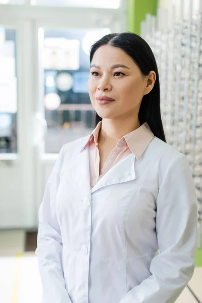 Asian ophthalmologist in white coat standing in optics shop on blurred background — стоковое фото