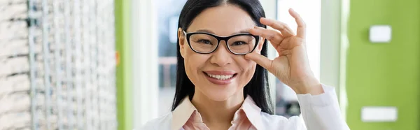 Joyful asian oculist adjusting eyeglasses while looking at camera in optics salon, banner - foto de stock