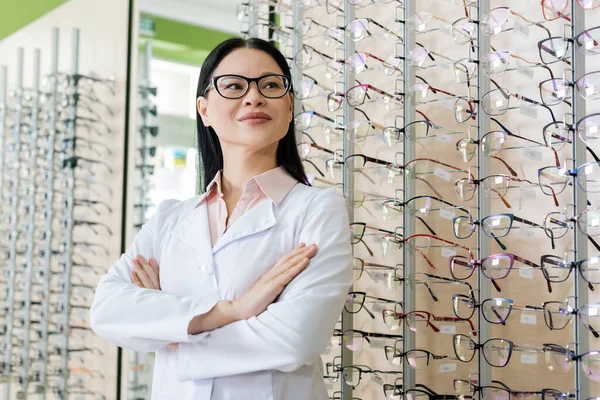 Pleased asian oculist standing with crossed arms near assortment of spectacles in optics shop — Photo de stock