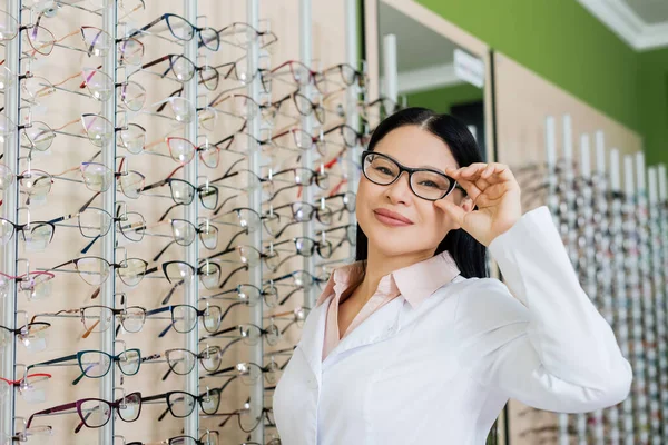 Pleased asian ophthalmologist adjusting eyeglasses while smiling at camera in optics salon — стоковое фото