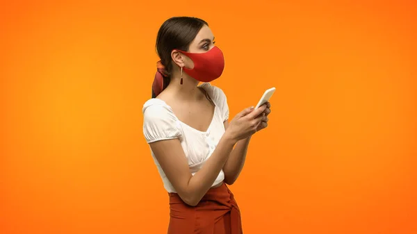Brunette woman in protective mask holding smartphone isolated on orange — Stock Photo