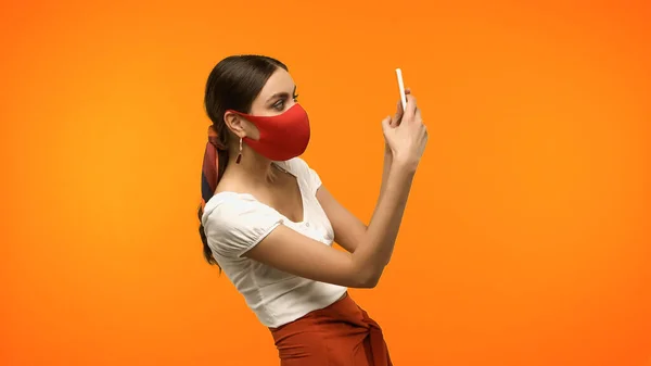 Young woman in protective mask taking photo on smartphone isolated on orange - foto de stock